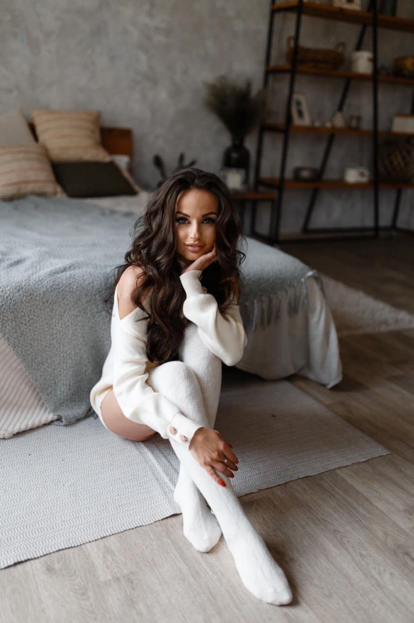 A woman sits on a bedroom rug wearing a white off-shoulder sweater and thigh-high socks, with a bed and shelves behind her.