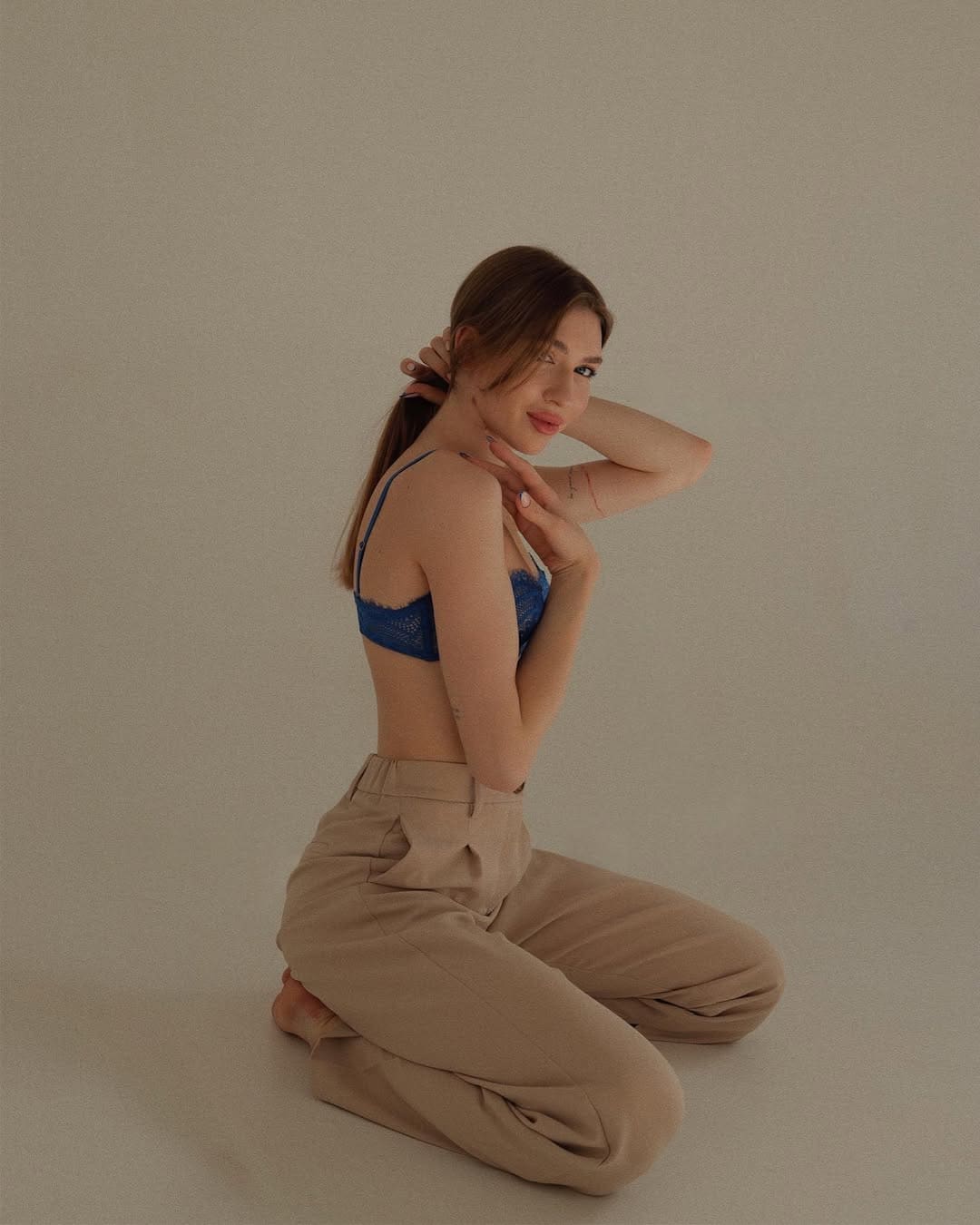 A woman with a low ponytail kneels on a plain floor in a blue lace bra and beige trousers, hands near her head, looking over her shoulder.