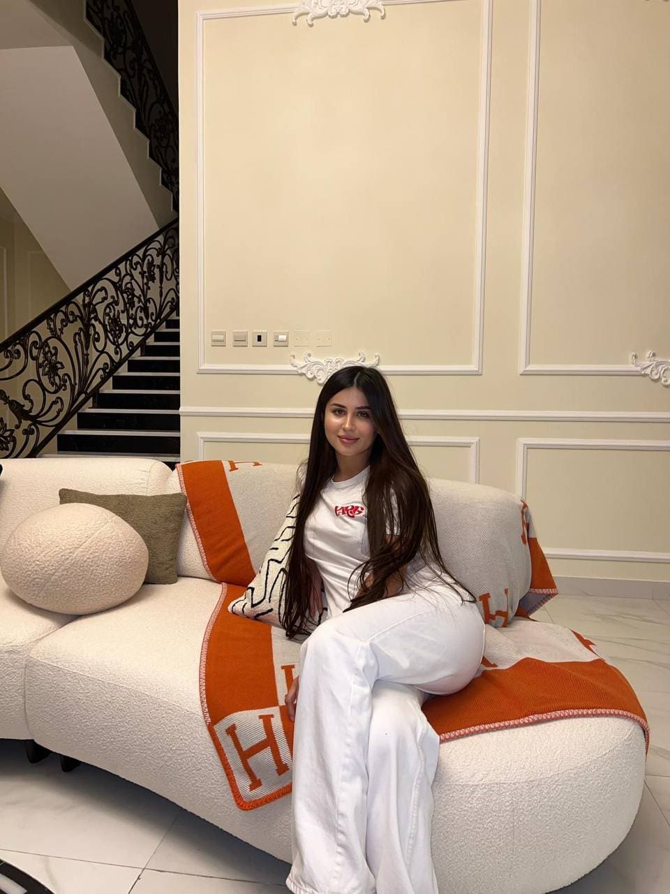 A woman in a white t-shirt and pants sits on a white sofa with an orange blanket, near cream paneled walls and a staircase with iron railing.