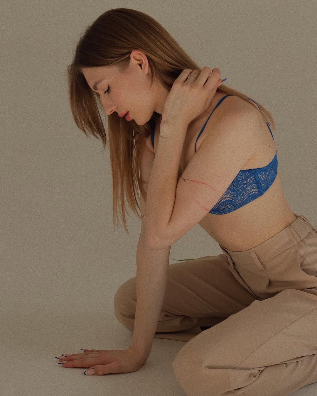 A woman with long light-brown hair kneels on a plain floor in a blue lace bra and beige trousers, one hand on the ground, the other on her neck.