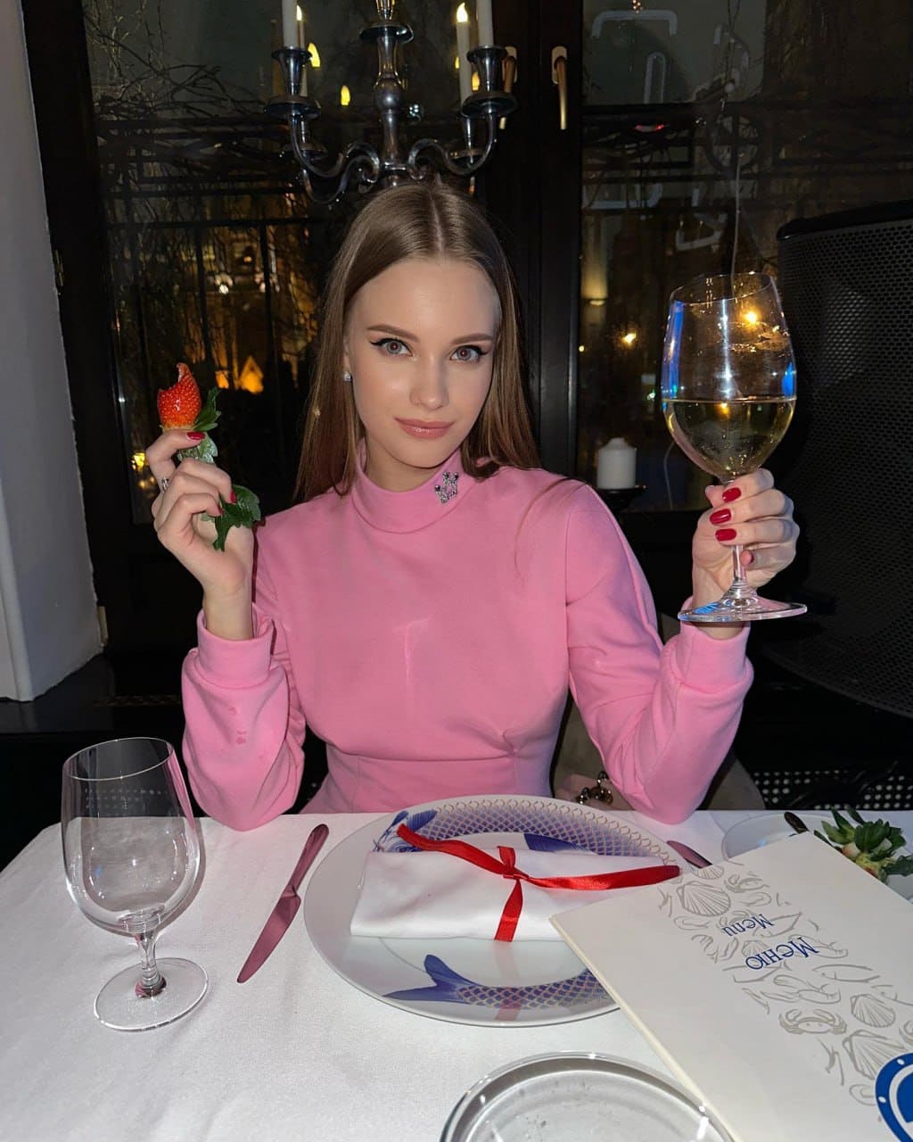 A woman in a pink turtleneck sits at a restaurant table, holding a wine glass and a strawberry.