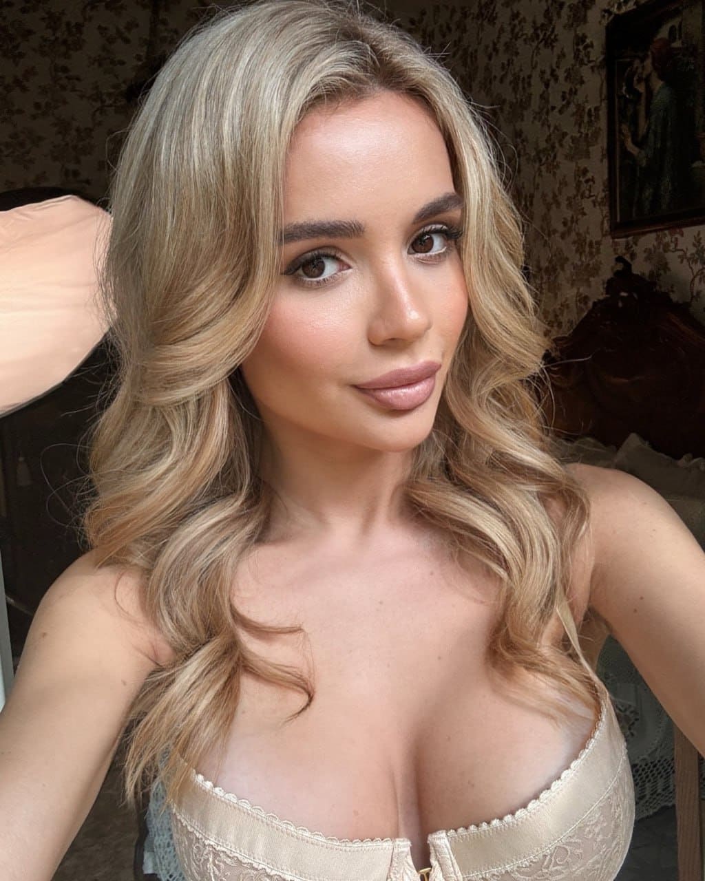 Sabina in a close-up selfie wearing a cream lace bustier, set against patterned wallpaper and a framed painting in a dim room.