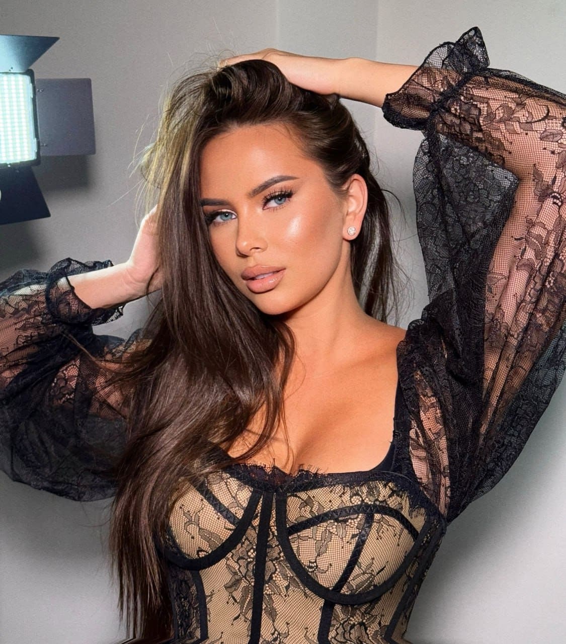 A woman with long dark hair poses with hands on her head, wearing a sheer black lace top, against a gray wall with a studio light.