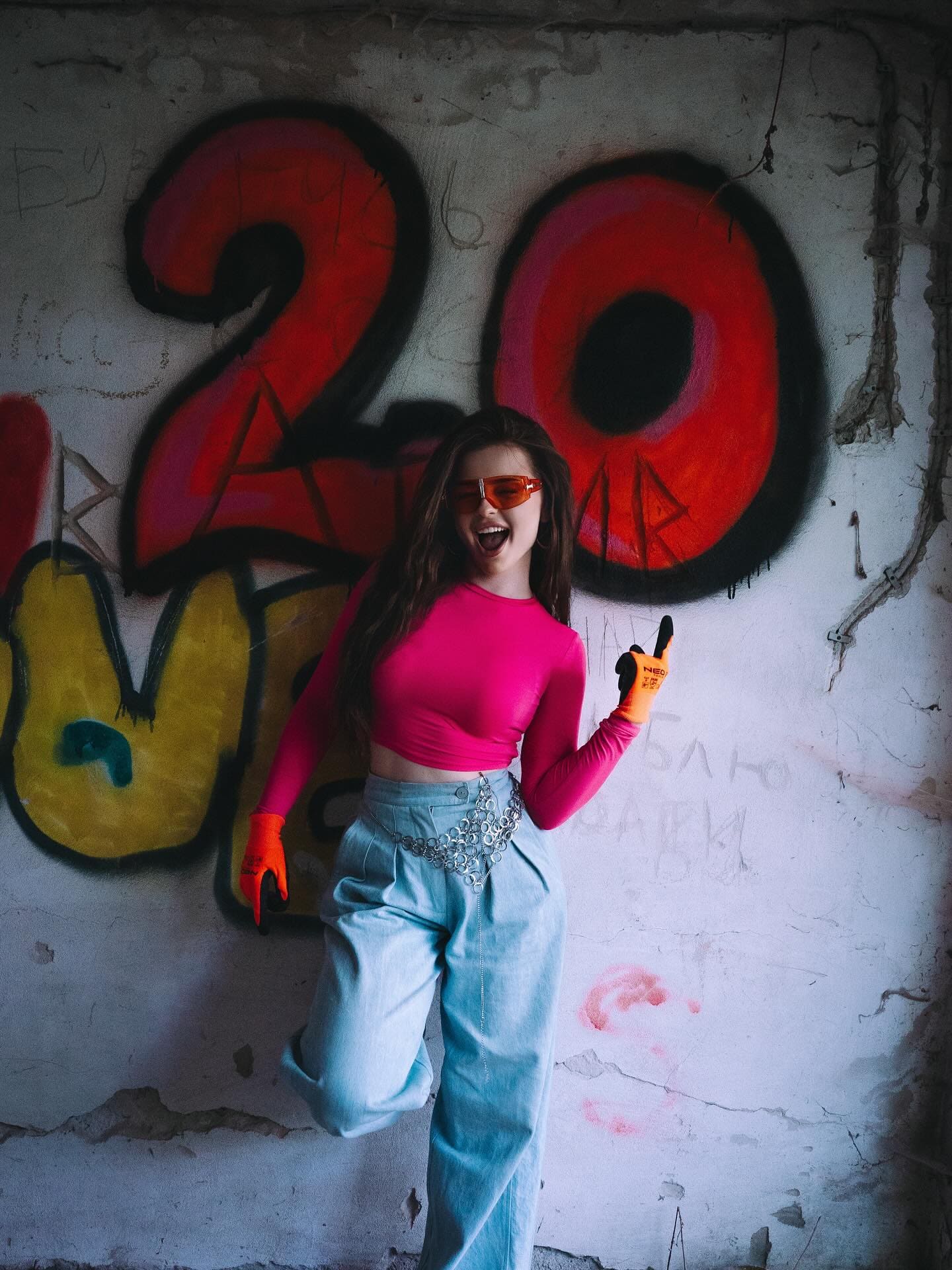 A woman in a pink crop top, blue wide-leg pants, and orange gloves poses playfully against a graffiti-covered concrete wall.