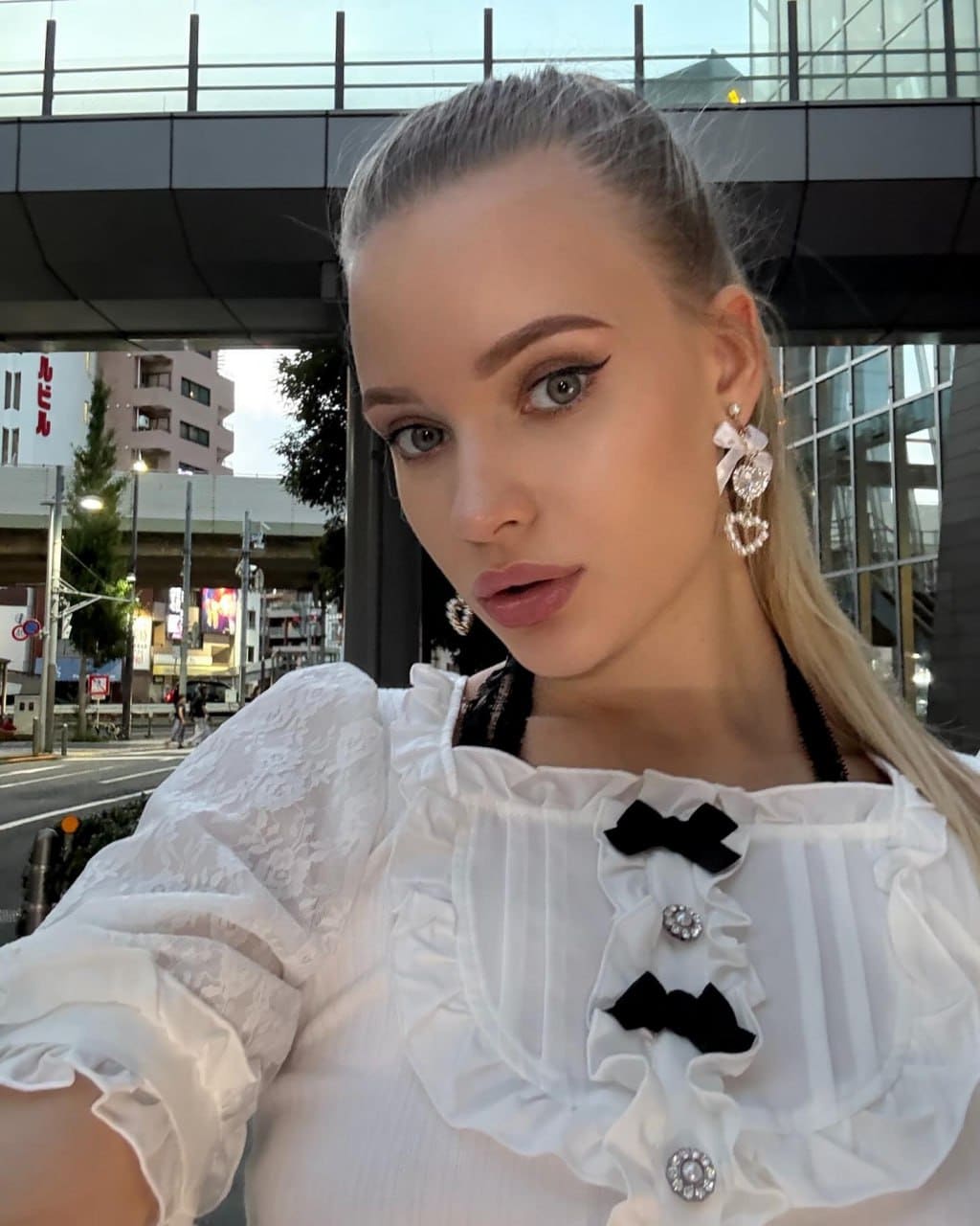 A blonde woman in a white ruffled blouse with black bows takes an outdoor selfie in an urban setting.