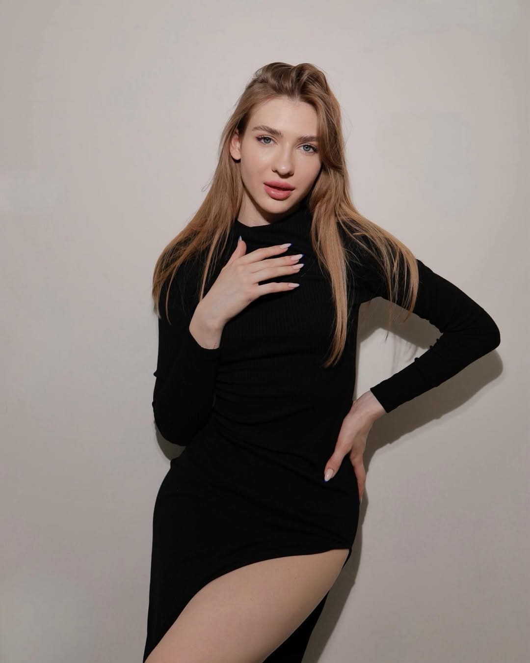 A woman with light-brown hair wears a black turtleneck dress with a thigh slit, resting a hand on her chest against a beige wall.