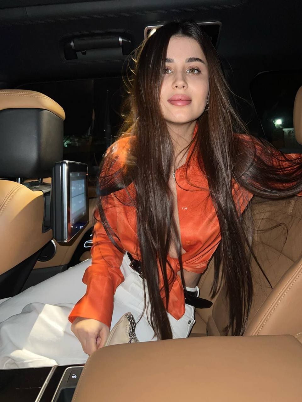A woman in an orange button-up shirt and white pants sits in a car's back seat, holding a small bag on her lap.
