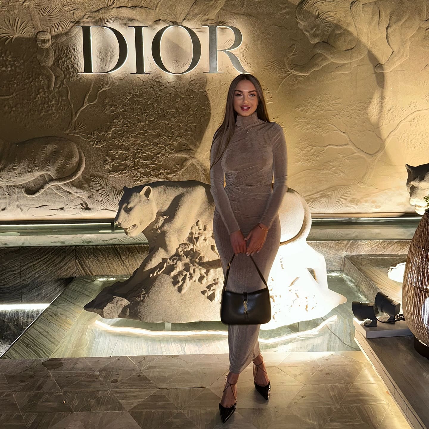A woman in a taupe turtleneck dress and black heels stands before a sculpted wall reading DIOR, holding a black handbag.