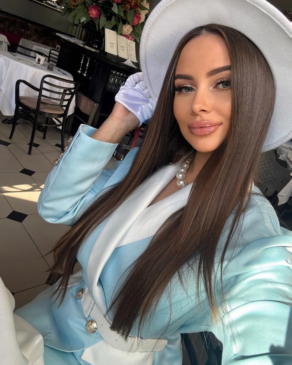 A woman with straight brown hair wears a white hat, blue and white jacket, and pearl necklace, posing in a restaurant with checkered floors.