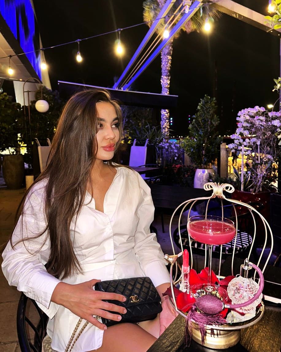 She sits at an outdoor night table in a white shirt, holding a black handbag, with a birdcage cocktail.