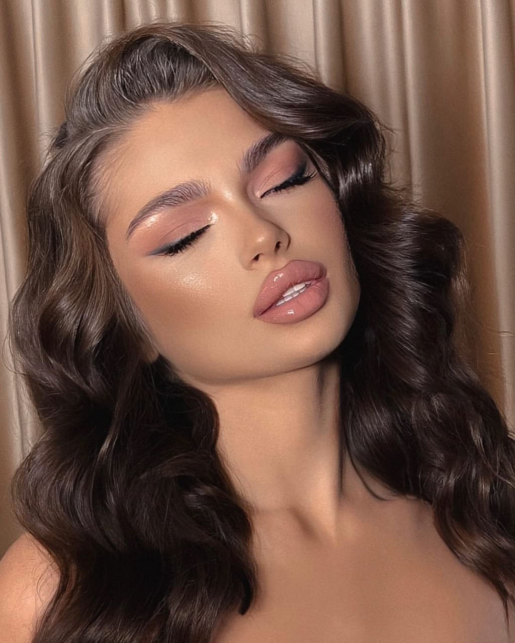 Close-up portrait of a woman with eyes closed, wearing pink eyeshadow and nude gloss, bare shoulders visible against a beige curtain.
