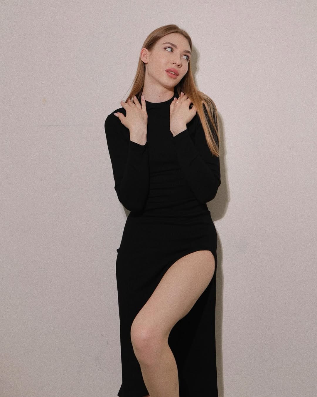 A blonde woman in a black long-sleeved dress with a slit raises her hands to her shoulders, one leg bent forward against a beige wall.