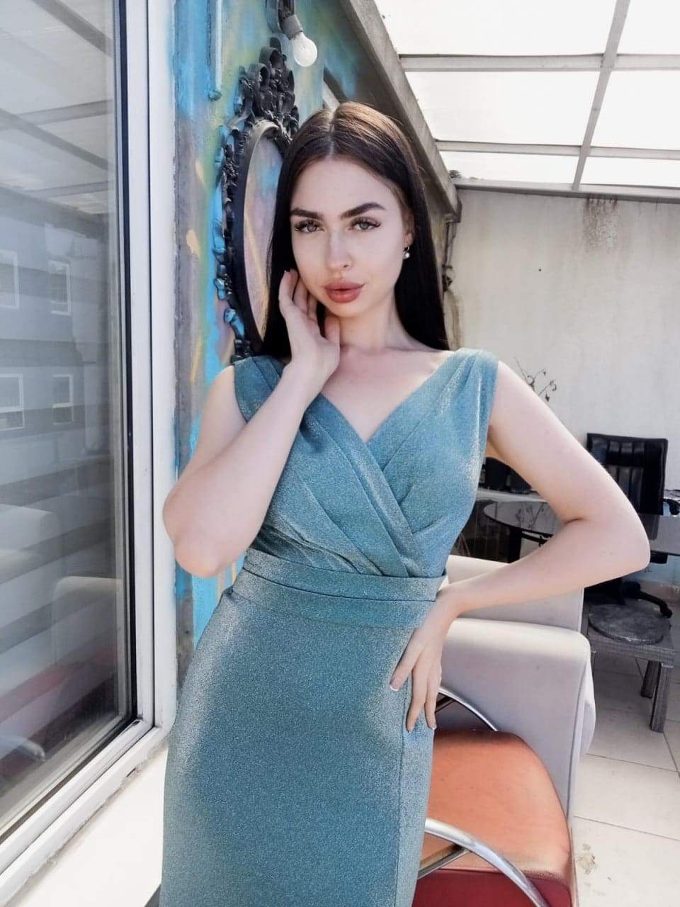 Clodia stands by a window in a teal sleeveless wrap dress, one hand on her cheek, with a chair and painted wall in the background.