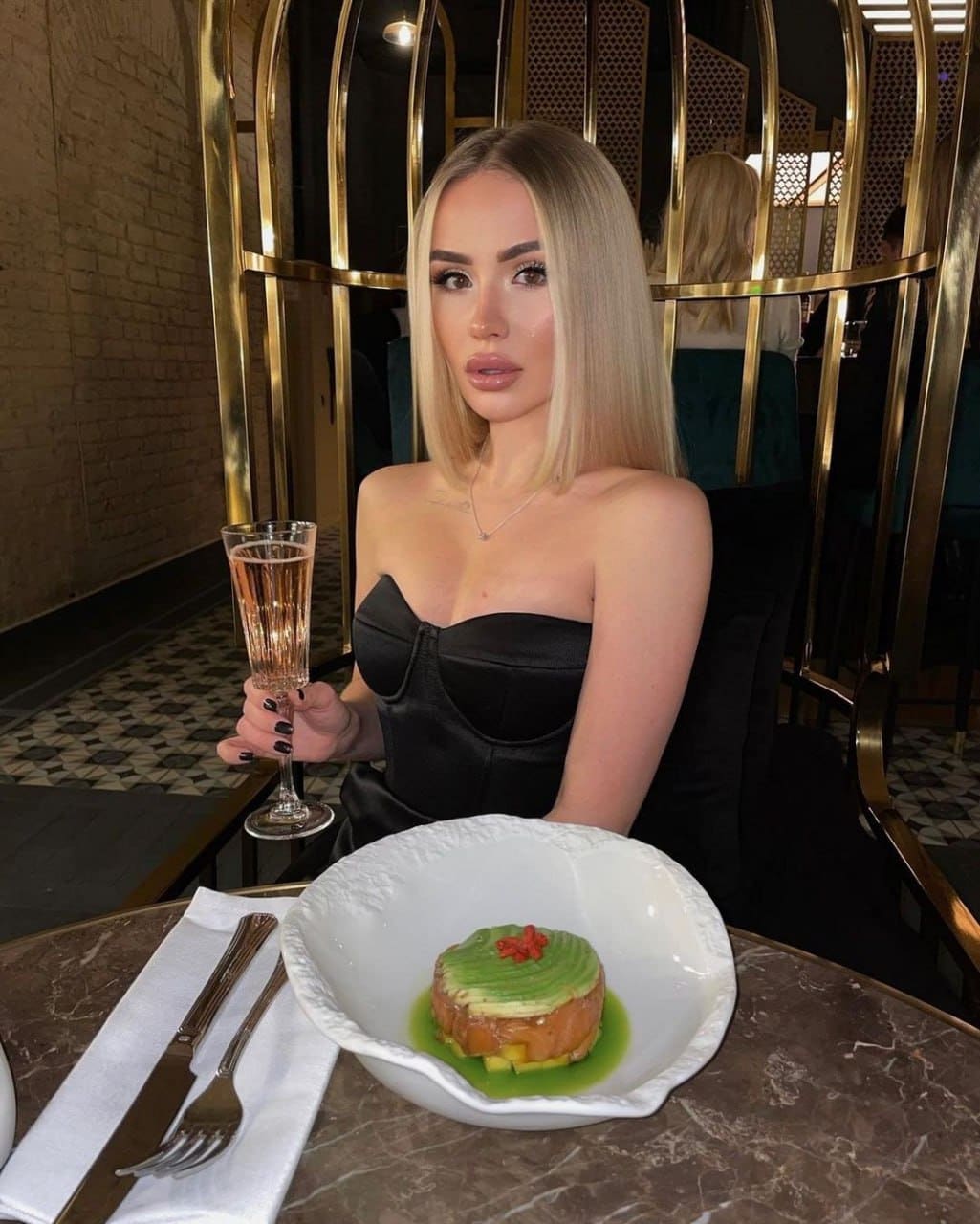 Sandra sits at a marble restaurant table in a black strapless corset top, holding a sparkling drink with dessert plated before her.