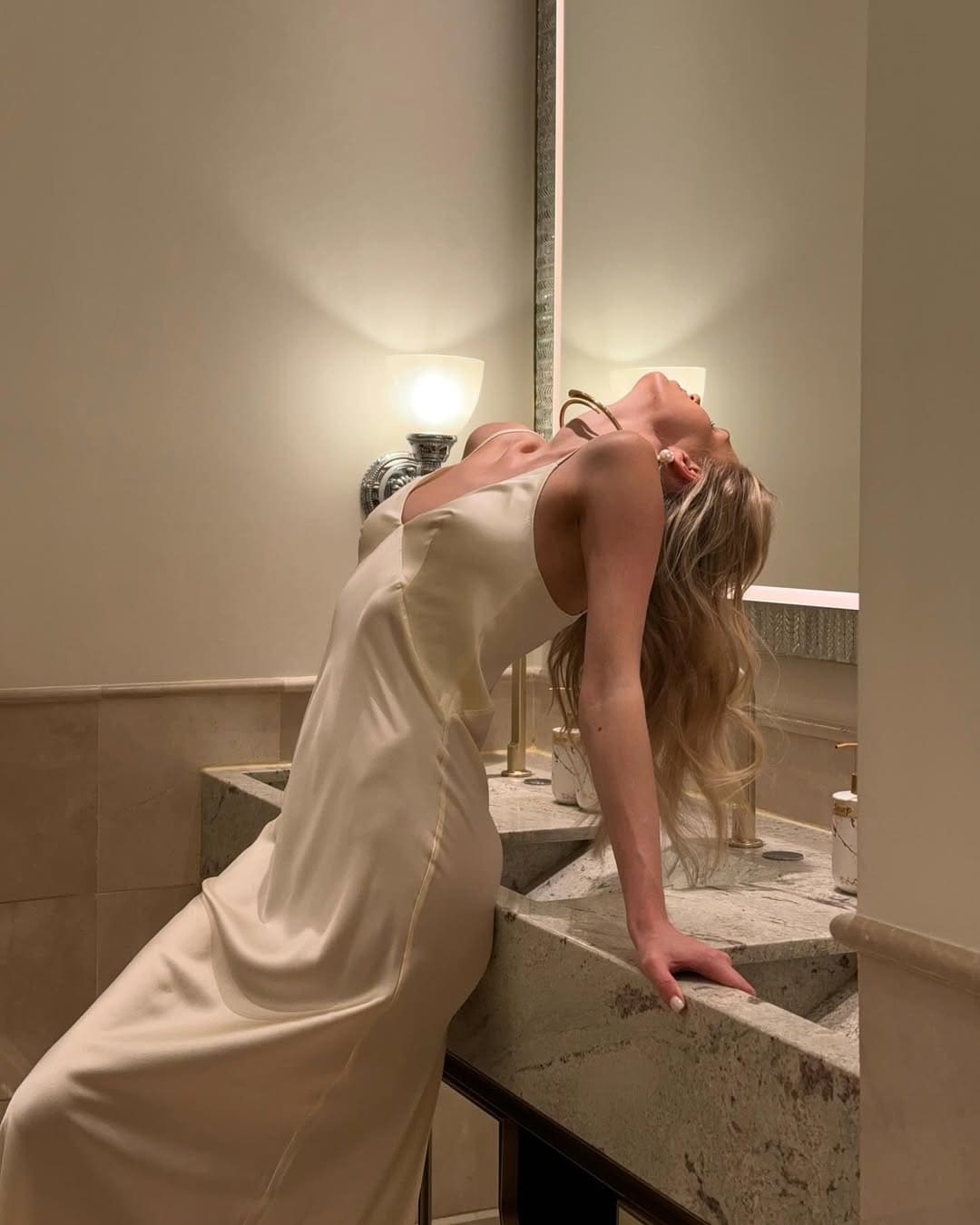 A woman in a cream satin slip dress leans back over a marble bathroom counter, head tilted up, one hand on the sink edge.