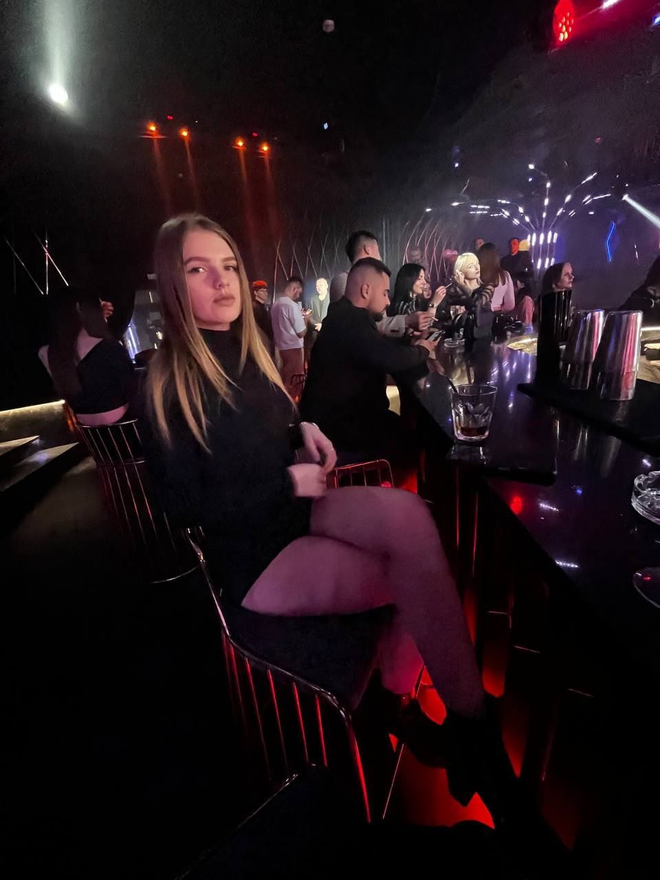 A woman in a black top sits on a metal chair at a dim club bar, with red lights and patrons behind.