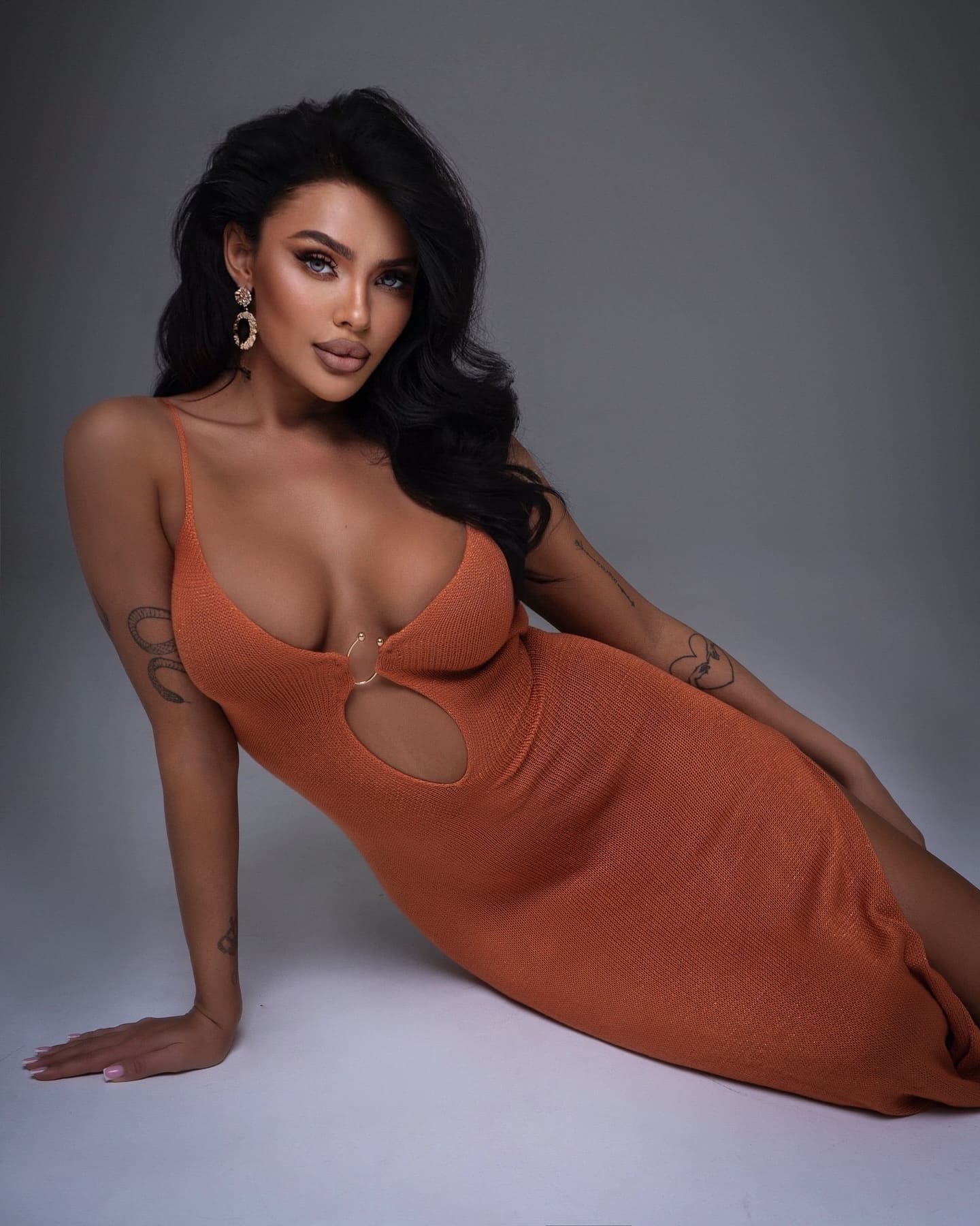 Alexia reclines on a white studio floor in an orange knit dress with cutouts, showing arm tattoos and hoop earrings.