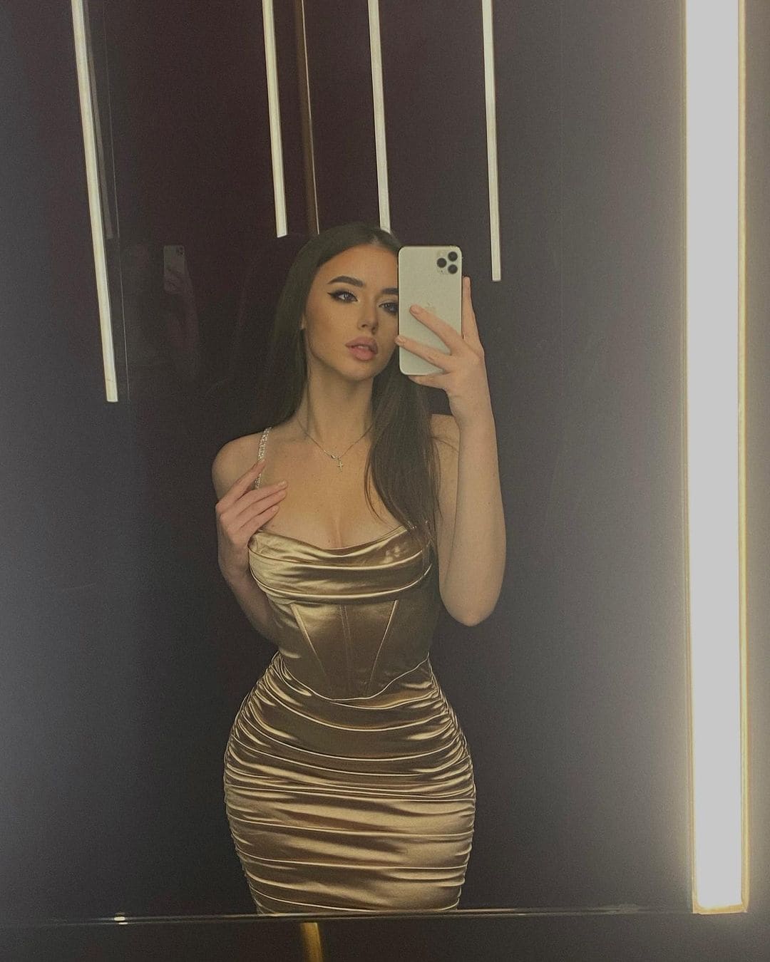 A mirror selfie shows a woman in a metallic gold satin dress with thin straps, standing against a dark wall with vertical light strips.