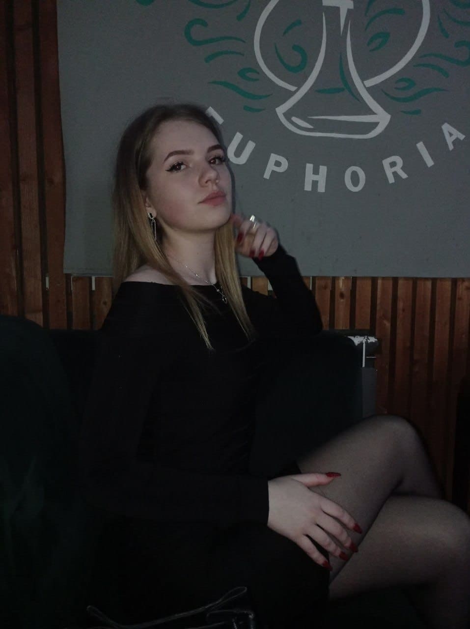 A blonde woman in an off-shoulder top sits on a dark couch, hand near chin, against a gray wall.