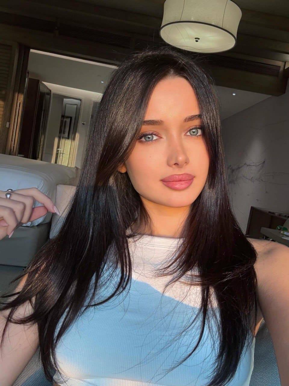 Close-up selfie of a woman with blue eyes in a hotel room, wearing a blue and white striped sleeveless top and a ring on her raised hand.