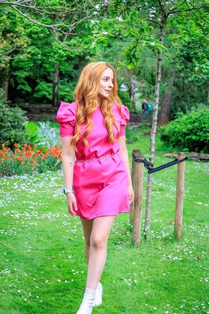 On a grassy lawn, she stands in a pink puff-sleeve dress with a heart belt and white boots, wearing a pearl headband, looking off to the side.
