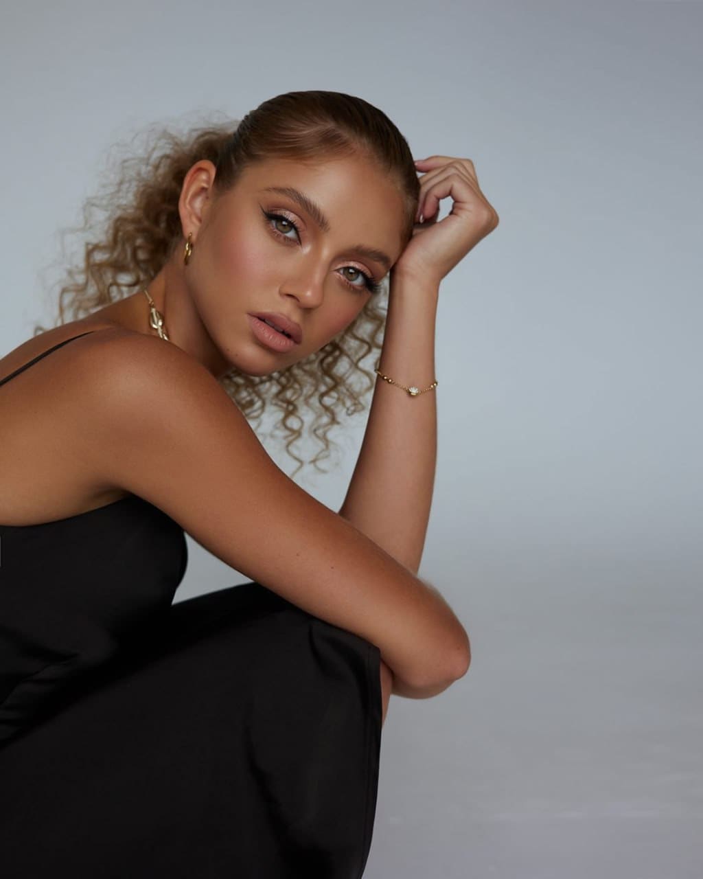 Luna sits in a black sleeveless dress against a gray studio background, one arm on her knee and the other raised to her head.
