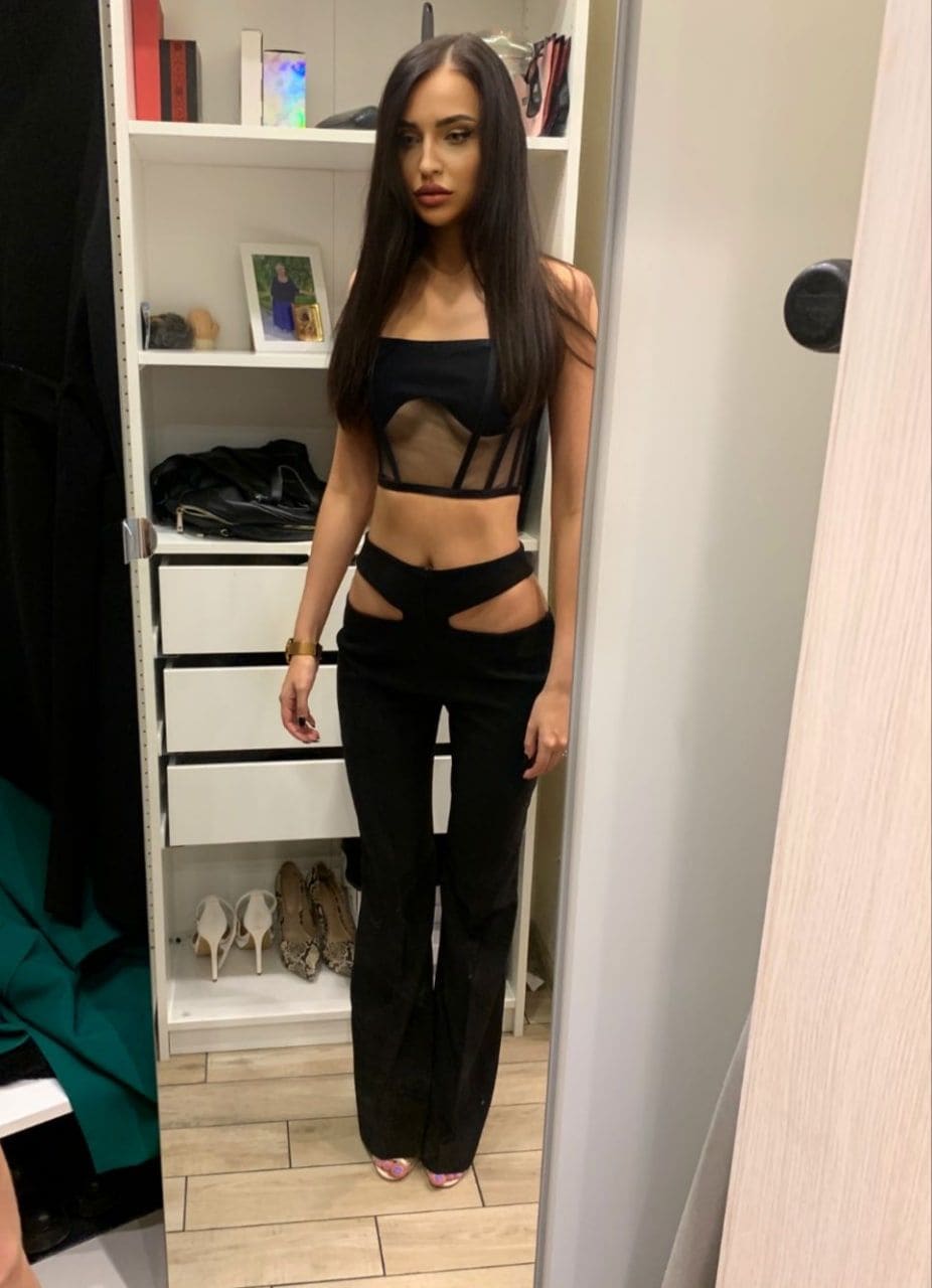 A woman stands in a doorway wearing a black sheer crop top and cut-out trousers, with a white shoe shelf behind her.