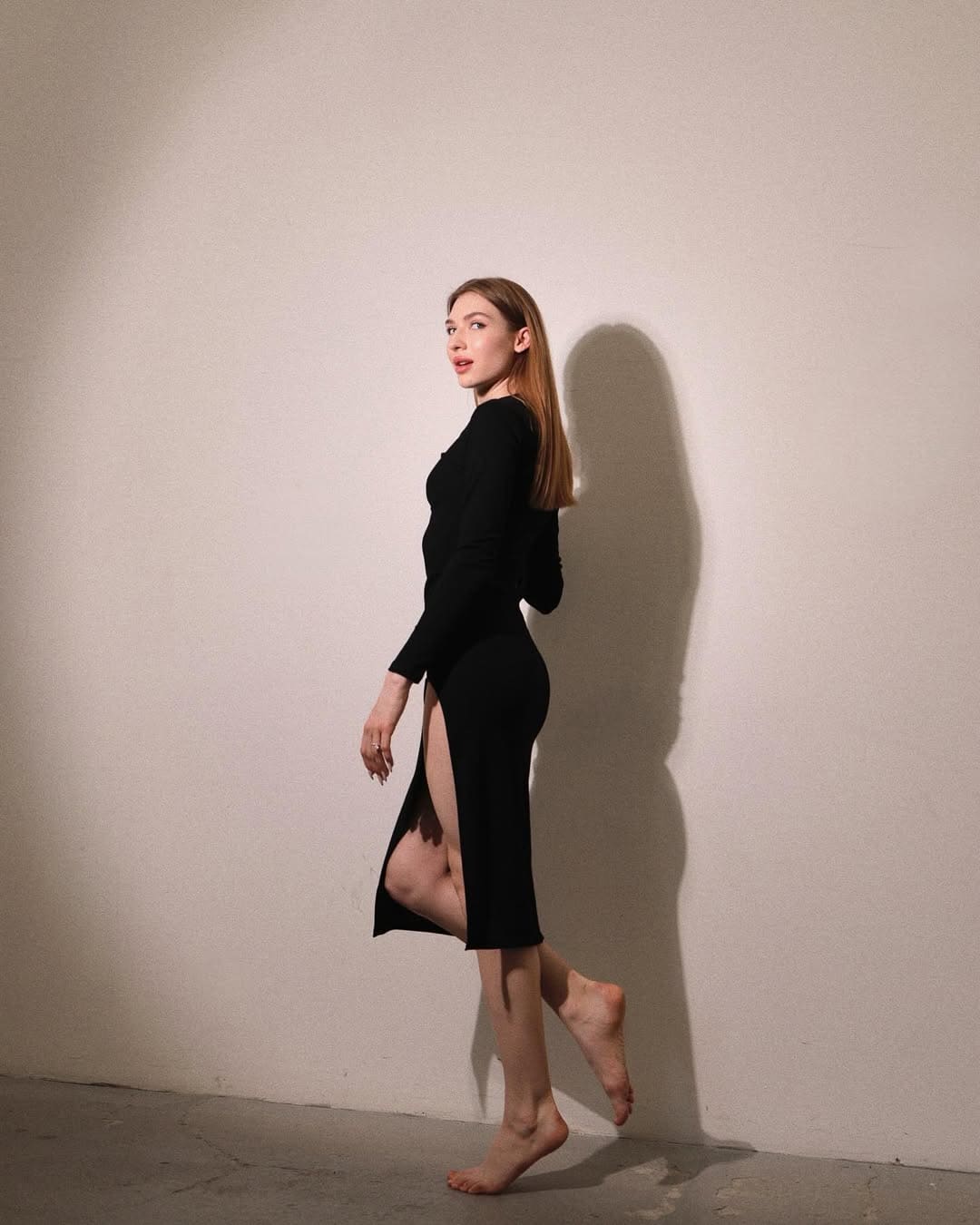 A barefoot woman with reddish-brown hair walks in profile against a white wall, wearing a black dress with a high side slit.
