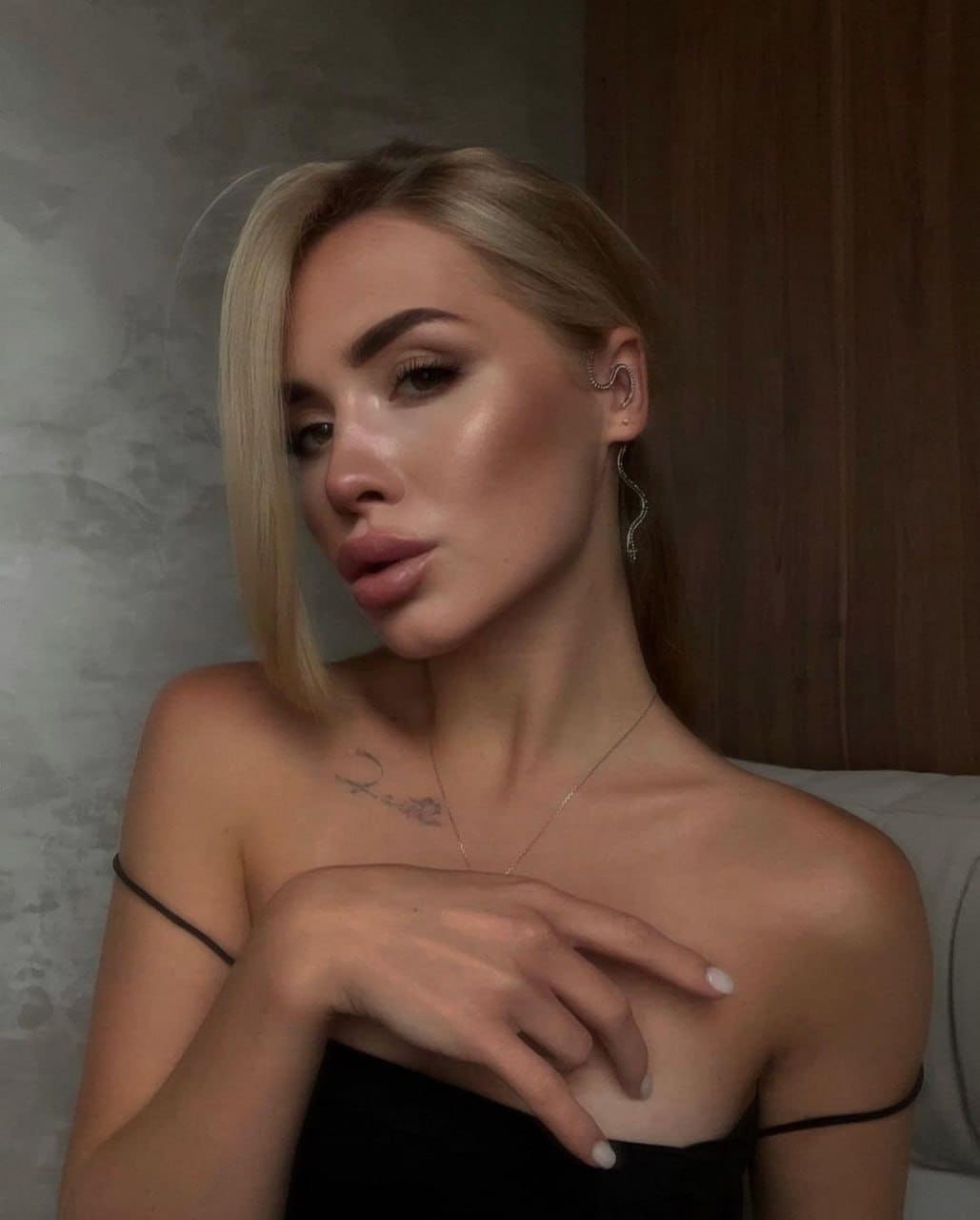 A blonde woman with a side-swept fringe poses in a black off-the-shoulder top, hand on chest, against a grey textured wall.