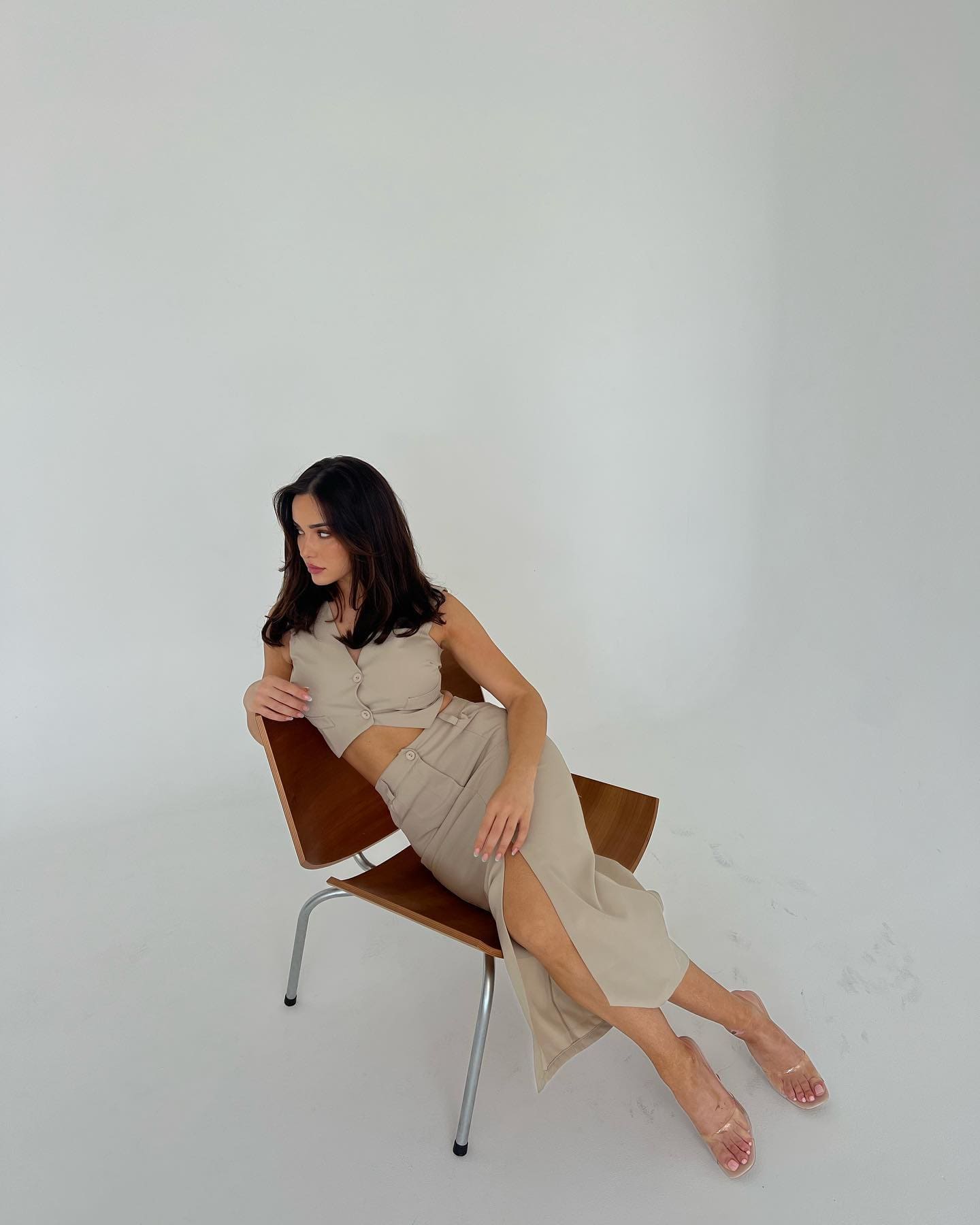 A woman sits sideways on a wooden chair wearing a beige crop top, slit skirt, and clear heels against a white background.
