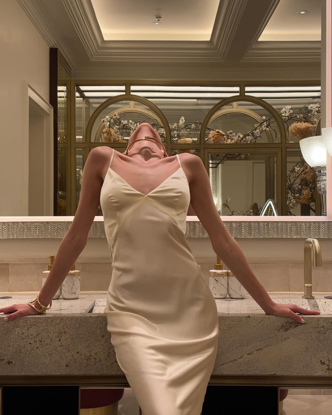 A dark-haired woman in a cream satin slip dress leans forward with head tilted back on a marble counter, in front of a mirror with arched gold frames.