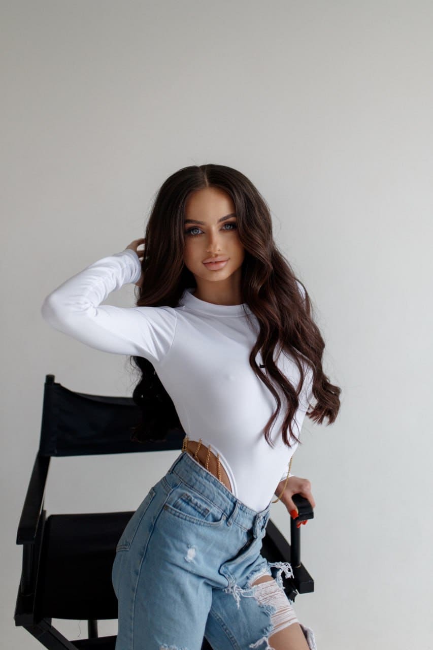 Leila stands by a black director's chair in a white cutout top and ripped jeans, one hand in her wavy hair against a light background.