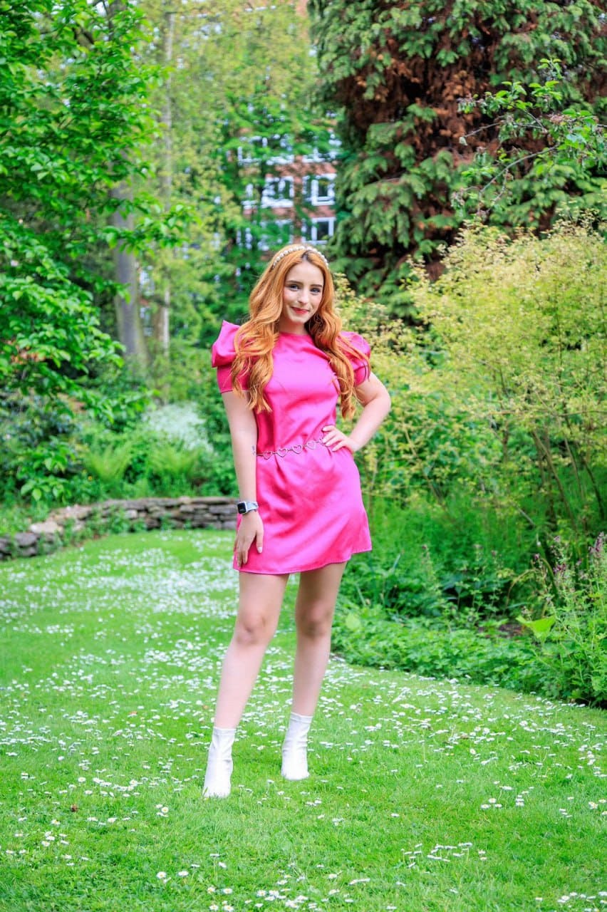 In a garden, she wears a bright pink dress with puffed sleeves and a heart belt, one hand on her hip, surrounded by green trees and shrubs.