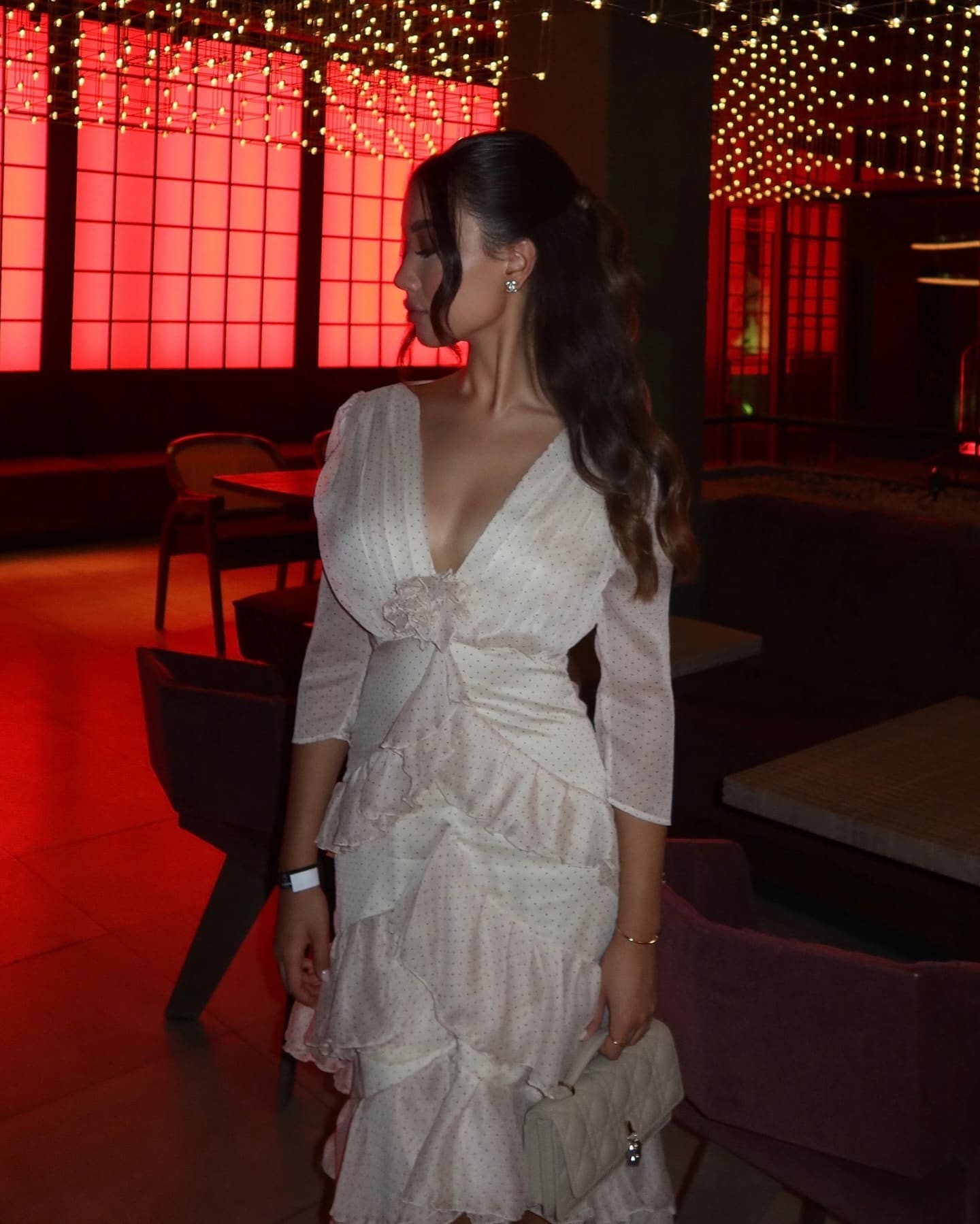 A woman in profile wears a white polka-dot dress and holds a quilted bag in a dim room with red-lit windows.