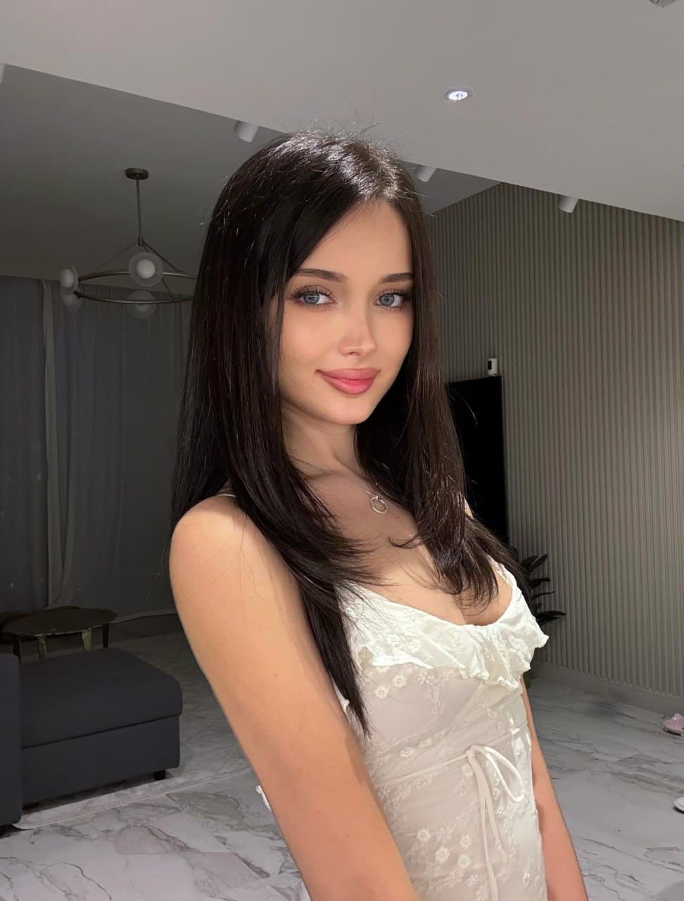 Woman standing in a room with marble floors, wearing a white lace dress with a ruffled neckline and a thin necklace, smiling slightly.