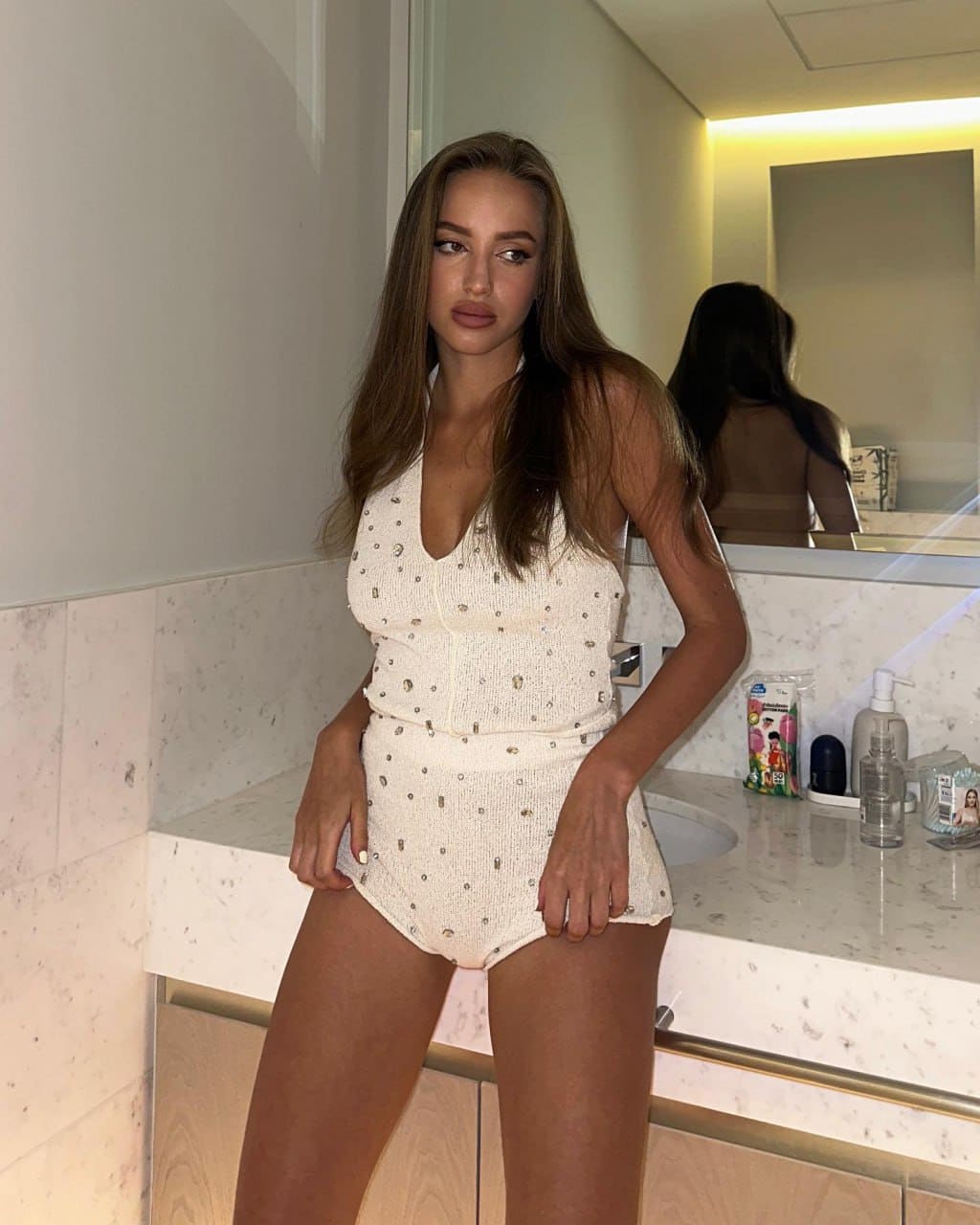 Anna stands in a bathroom wearing a white studded one-piece swimsuit, posing with hands on hips in front of a marble counter and mirror.