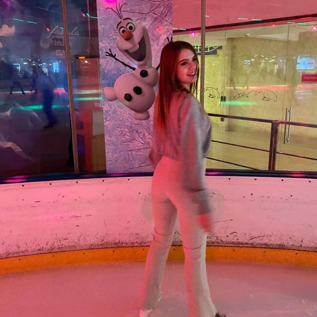 A woman in grey loungewear stands on an ice rink near a glass wall with an Olaf figure and pink light.