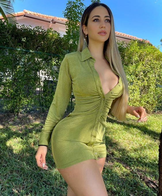In a garden, a woman stands wearing a tight olive-green long-sleeved mini dress with an open neckline, one hand on her hip.