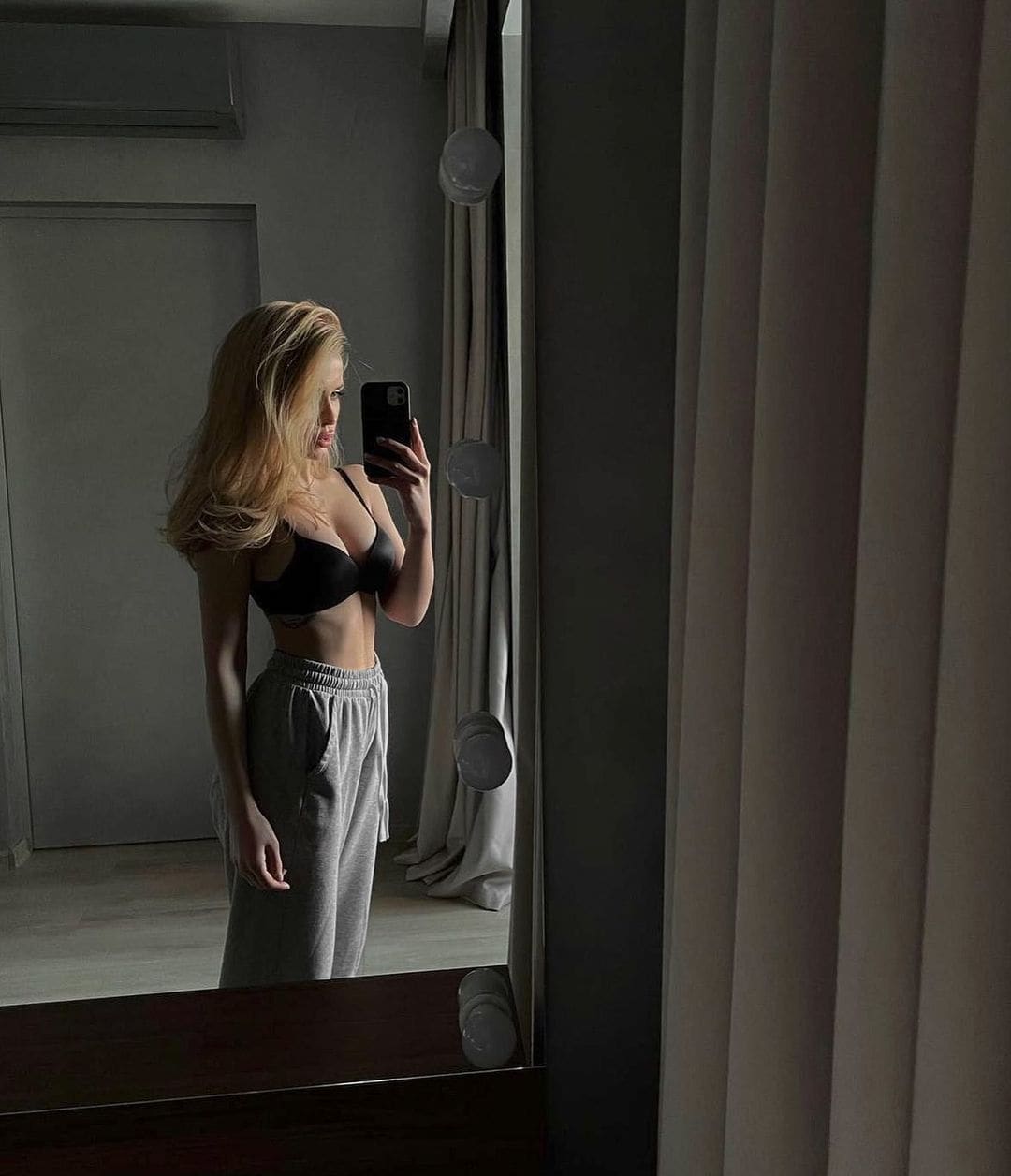 A blonde woman takes a mirror selfie in profile, wearing a black bra and grey sweatpants in a dimly lit room with curtains.