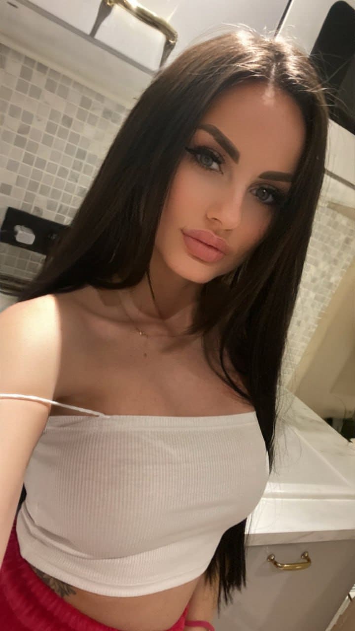A woman poses in a bathroom wearing a white crop top with a slipping strap, showing a small midriff tattoo against a tiled wall.