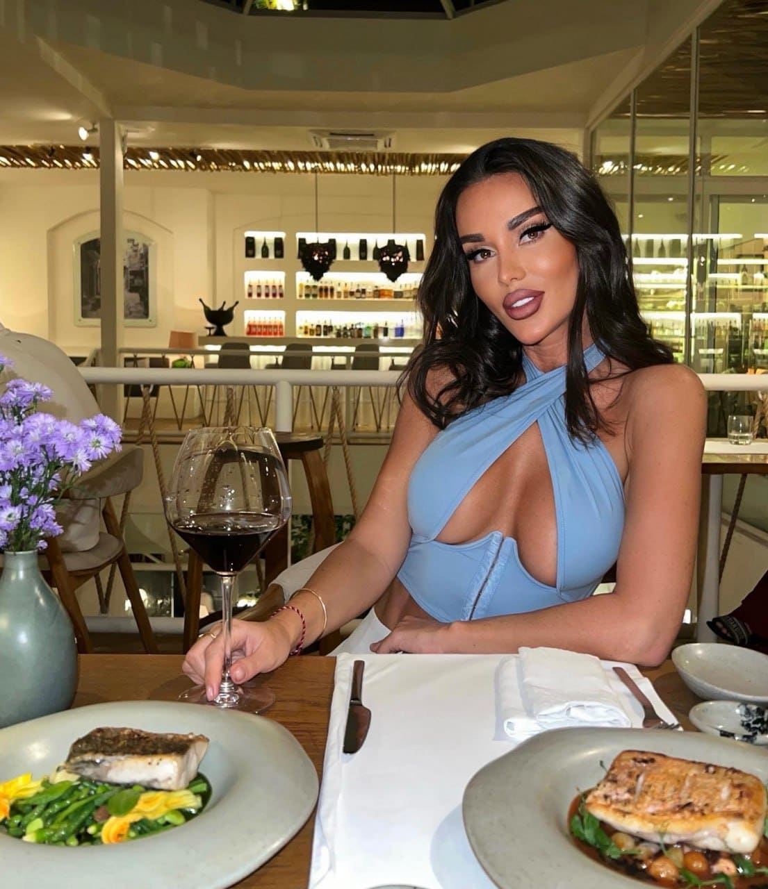 Yulia sits at a restaurant table in a light blue halter top and white bottoms, holding red wine, with food and purple flowers nearby.