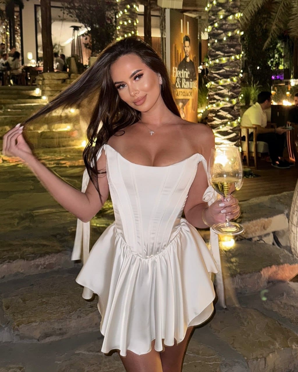 A woman in a white off-shoulder mini dress stands outdoors at night, holding a glass of white wine near string lights and a stone patio.