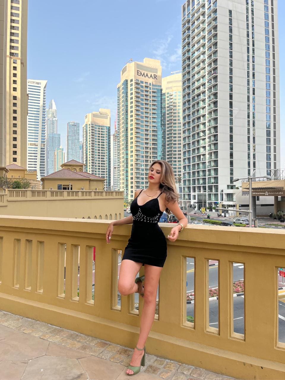 Alexa stands on a stone terrace in a short black dress with a beaded chest and green heels, leaning against a yellow balustrade with a city skyline behind.