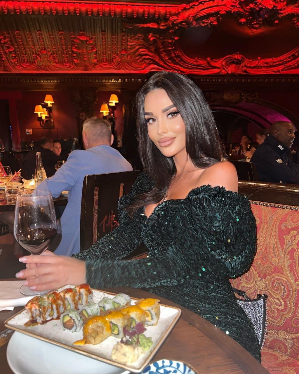 A woman in a black off-the-shoulder dress sits at a table with sushi and red wine, illuminated by warm red ambient lighting.