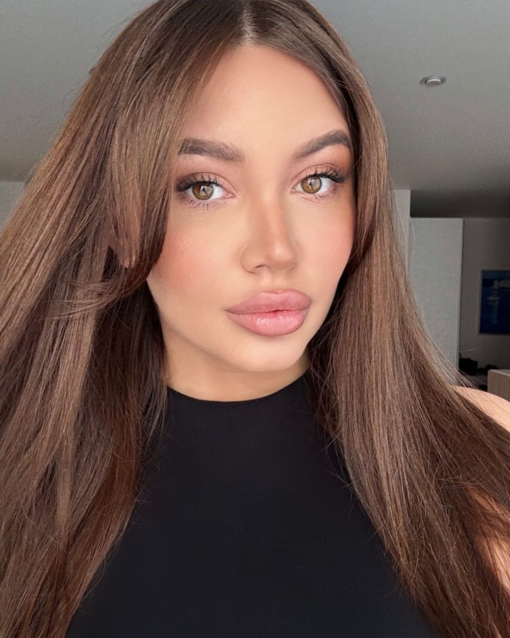 Close-up portrait of a woman with long straight brown hair and a black high-neck top, set against a plain indoor background with a ceiling light.