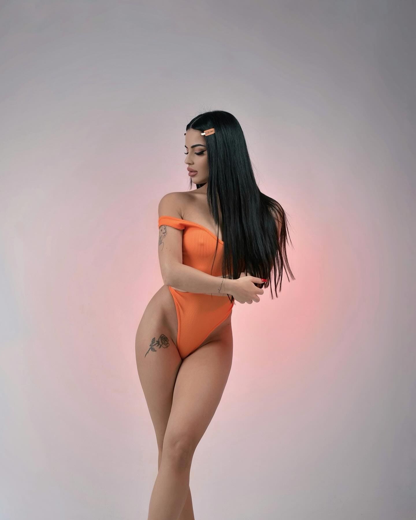 Profile view of a woman in an orange off-the-shoulder swimsuit, showing a rose tattoo on her hip against a pink-lit background.