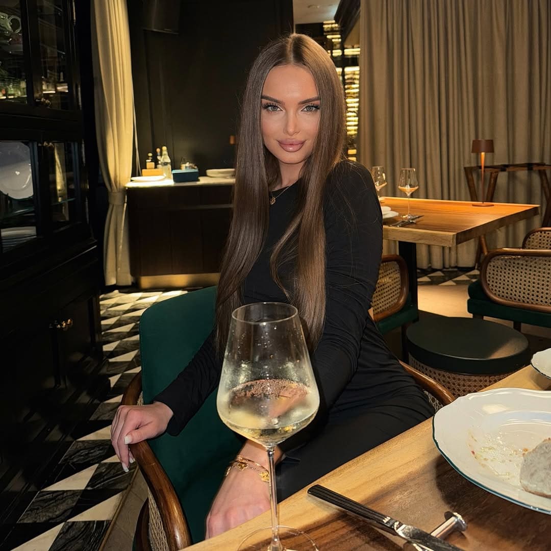 Inna sits at a wooden restaurant table in a fitted black top, with a glass of white wine in the foreground and dark cabinetry behind.