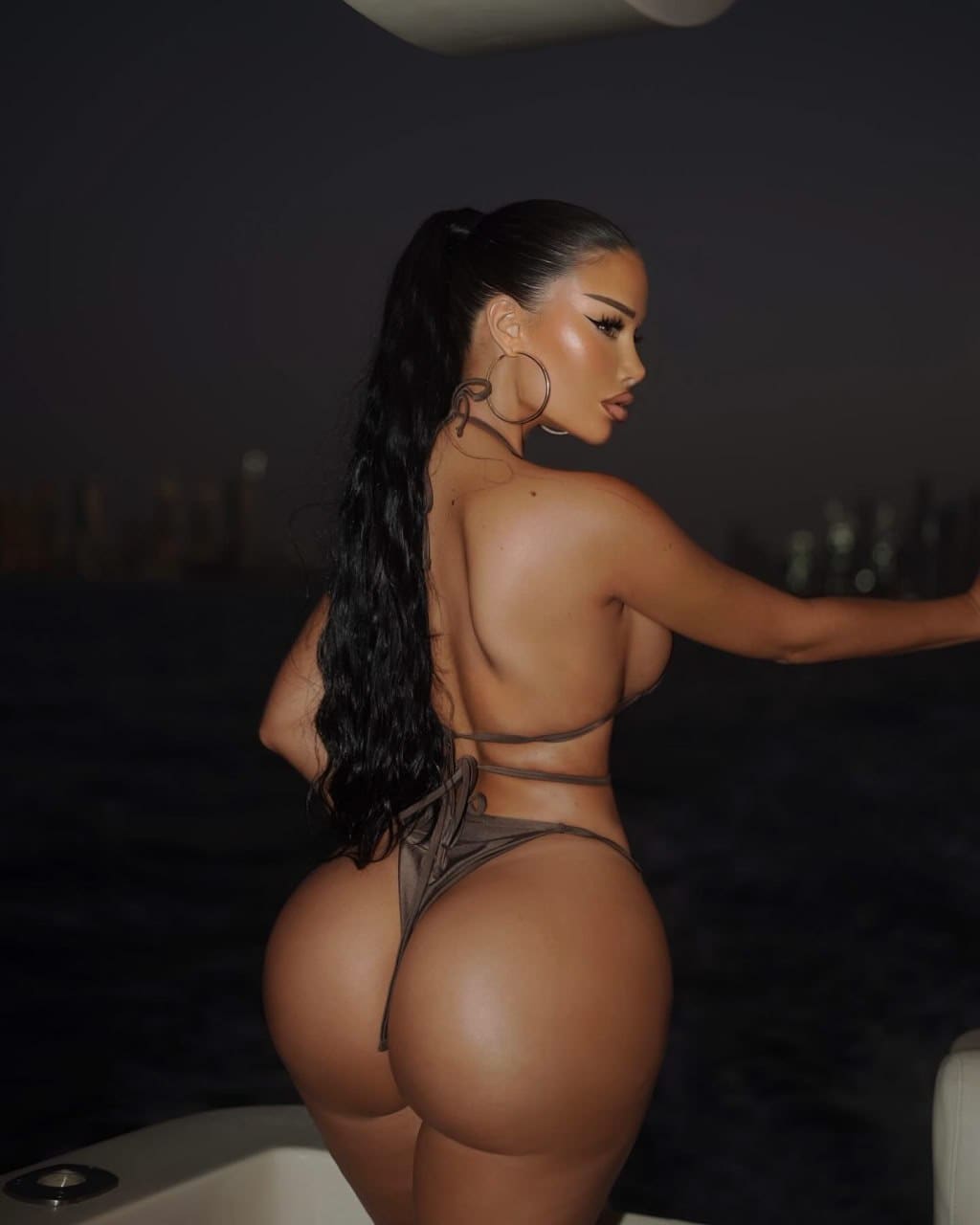 A woman with a high ponytail stands in profile on a boat at night, wearing a strappy brown bikini against a dark skyline.