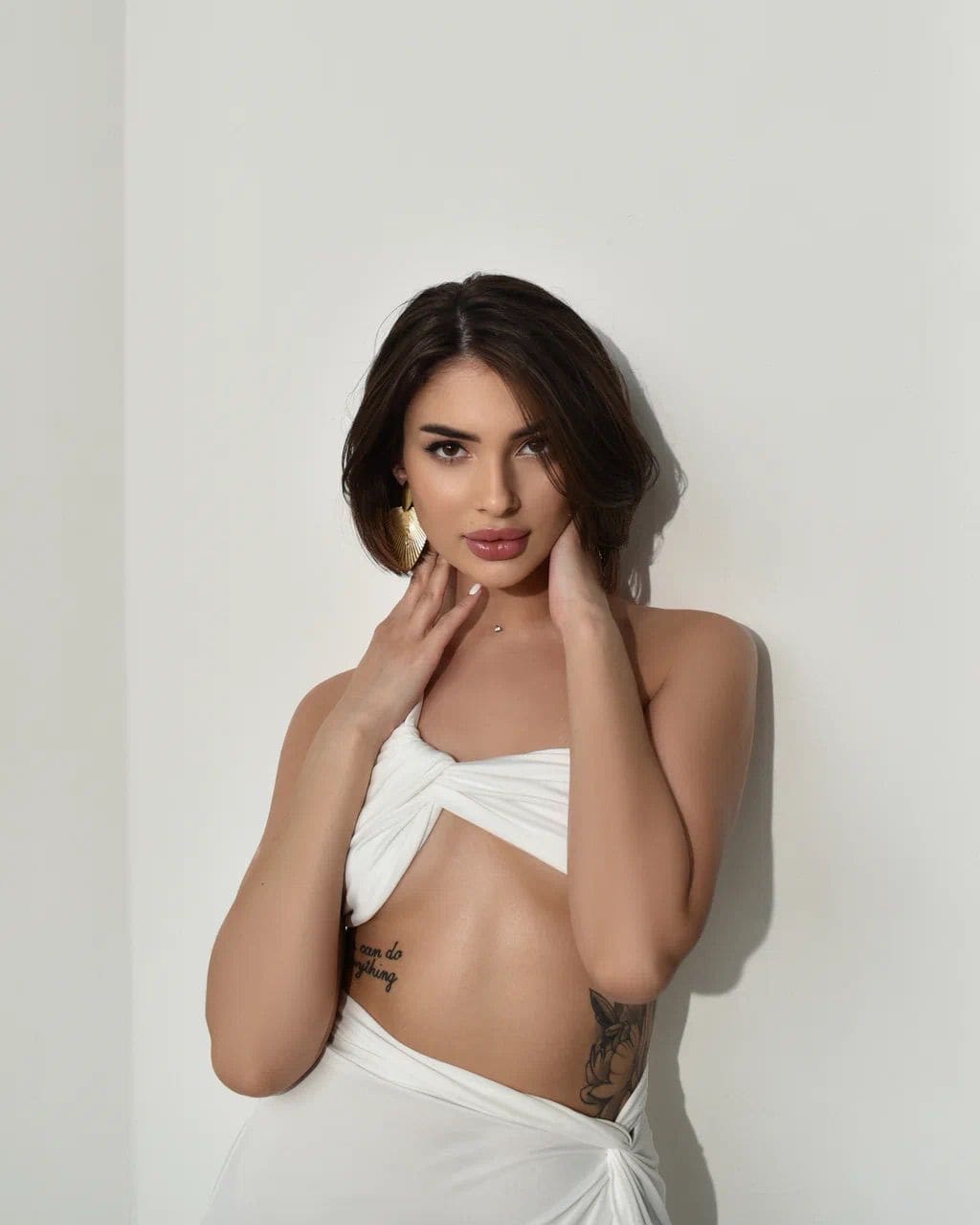 Alla stands against a white wall in a white bandeau and wrap skirt, framing her face with raised hands, revealing midriff tattoos.