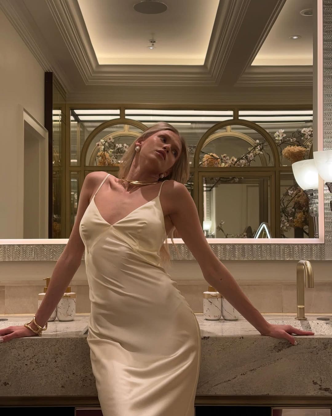 A blonde woman in a cream satin dress and gold jewelry leans on a marble counter, with a mirror reflecting arched windows and flowers behind her.